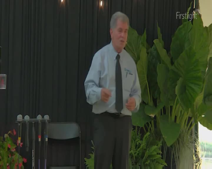 A man in a light blue shirt and dark tie gestures with his hands as he speaks. He stands before a backdrop of dark curtains, flanked by lush green foliage. A man in a light blue shirt and dark tie gestures with his hands as he speaks. He stands before a backdrop of dark curtains, flanked by lush green foliage.