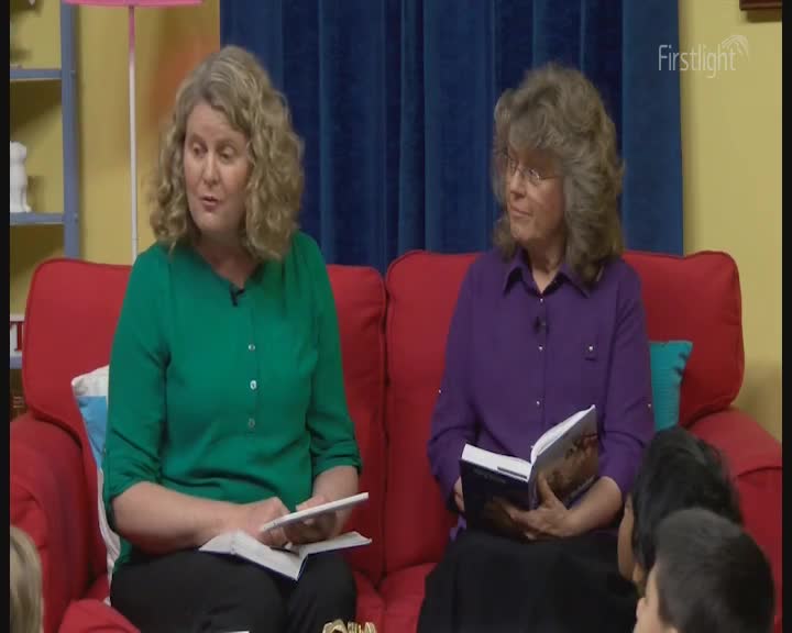 Two women sit on a bright red couch, one in a green top, the other in purple, holding books. The woman in purple turns a page, her gaze directed towards a group of children gathered before them. Two women sit on a bright red couch, one in a green top, the other in purple, holding books. The woman in purple turns a page, her gaze directed towards a group of children gathered before them.