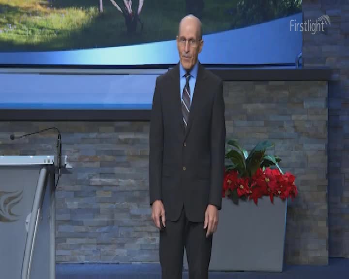 A man in a dark suit stands on a stage, facing forward. Behind him, a large screen displays a pastoral scene, and a potted plant with vibrant red poinsettias sits to his right. A man in a dark suit stands on a stage, facing forward. Behind him, a large screen displays a pastoral scene, and a potted plant with vibrant red poinsettias sits to his right.