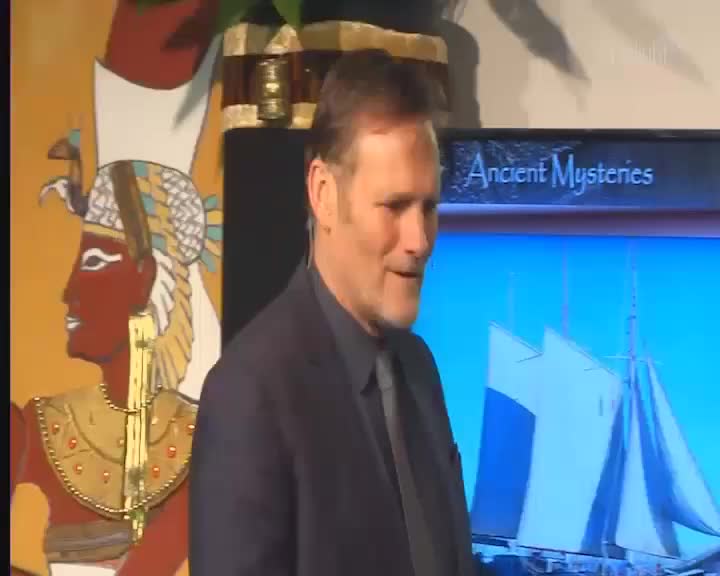 The man in the dark suit smiles as he stands beside a vibrant mural of an ancient Egyptian figure. Behind him, a screen displays the words "Ancient Mysteries" above a bright blue image of a sailing ship. The man in the dark suit smiles as he stands beside a vibrant mural of an ancient Egyptian figure. Behind him, a screen displays the words "Ancient Mysteries" above a bright blue image of a sailing ship.