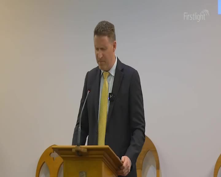 A man in a dark suit and yellow tie stands at a wooden lectern, speaking into a microphone. The Firstlight logo appears in the upper right corner of the screen.