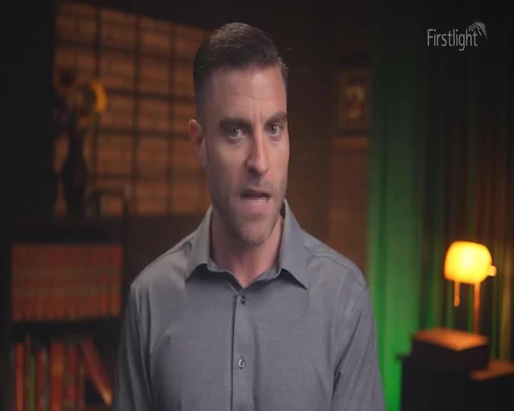 A man in a grey shirt speaks directly to the camera. Behind him, shelves filled with books line a wall, and a warm lamp casts a glow.