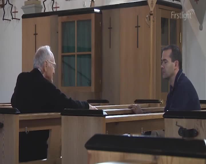 An older man in a black suit leans forward, his hand resting on the pew. Across from him, a younger man sits with his eyes closed, his own hand on the wood. The light filtering through the church windows casts a quiet glow.