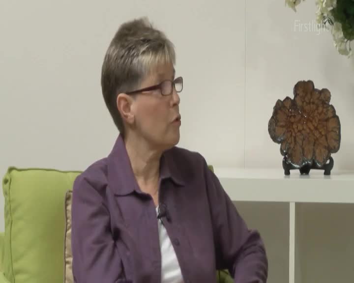 A woman with short grey hair and glasses speaks, her purple jacket open over a white shirt. Beside her, a textured, brown object sits on a dark stand.