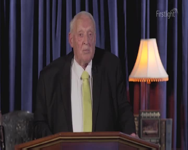 An older gentleman in a suit and bright yellow tie stands behind a wooden lectern. He looks directly ahead, his expression serious.