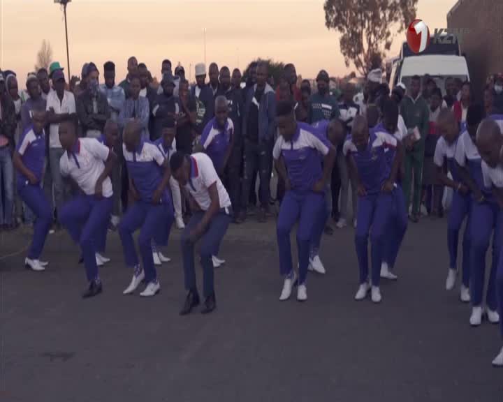A group of young men in blue and white uniforms are performing a synchronized dance, their knees bent low as they move in unison. A crowd of onlookers watches from the side of the street, some holding up phones, as the 1KZN TV logo appears on a nearby vehicle. A group of young men in blue and white uniforms are performing a synchronized dance, their knees bent low as they move in unison. A crowd of onlookers watches from the side of the street, some holding up phones, as the 1KZN TV logo appears on a nearby vehicle.