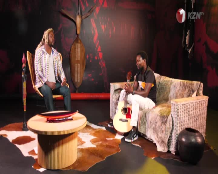Two men sit in a studio setting, one on a chair holding a guitar, the other on a couch with his guitar resting on his lap. The backdrop features traditional South African art and the 1KZN TV logo.