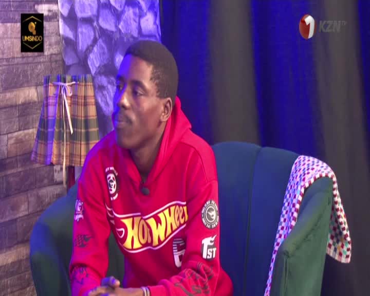 A young man in a bright red hoodie, emblazoned with a "Hot Wheels" logo, sits on a plush green chair. He's on a set for 1KZN TV, the KZN province's local broadcaster.