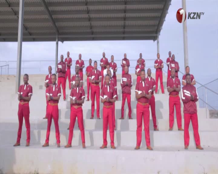 A group of men in matching maroon and white tracksuits stand on stadium bleachers. They are all facing forward, hands clasped in front of them.