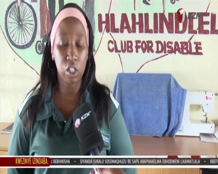 A woman speaks into a KZN microphone, her words captured for broadcast. Behind her, the wall displays "HLAHHLINDLE CLUB FOR DISABLE" in bold red letters.