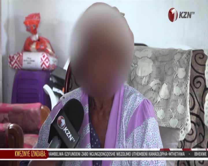 A KZN TV reporter holds a microphone up to a woman sitting in a room. The woman is wearing a patterned robe, and her face is blurred.