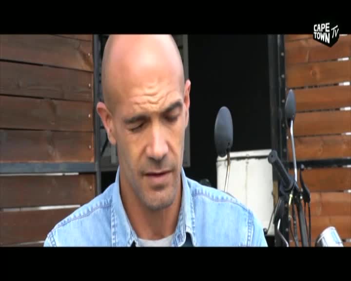 A bald man in a denim shirt looks down, his brow furrowed. Behind him, a motorcycle's handlebars and mirrors are visible against a wooden backdrop, with the Cape Town TV logo in the corner.