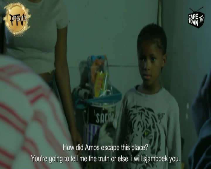 A young boy stands looking directly ahead, his expression serious. Behind him, a woman in a white top is partially visible, and a cluttered shelf holds various items.