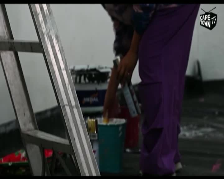 A person in bright purple trousers dips a brush into a teal paint bucket. The Cape Town TV logo is visible in the upper right corner.
