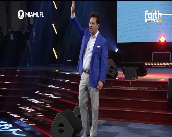 A man in a blue suit and grey trousers stands on a stage, his right arm raised. The Faith TV logo is visible on a screen behind him, alongside a phone number for South Africa.