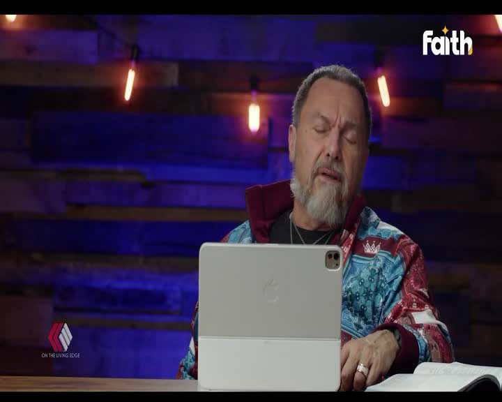 A man with a grey beard, wearing a vibrant jacket, speaks with his eyes closed. He's seated at a table with an open iPad and a Bible, likely for a Faith TV broadcast from South Africa.