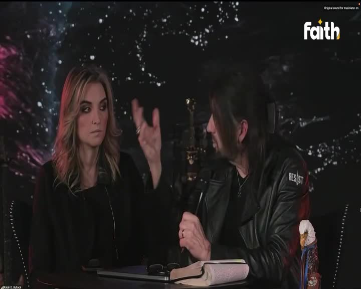 A woman with blonde hair gestures with her hand as she speaks, while a man with dark hair, wearing a leather jacket with "RESIST" on it, listens intently. They are seated at a table with a Bible and glasses, likely for a broadcast on Faith TV in South Africa.
