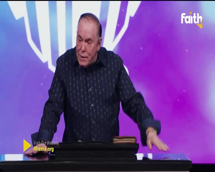 The speaker gestures with his right hand, his fingers splayed over a blue surface. A wallet rests on a dark stand to his left.