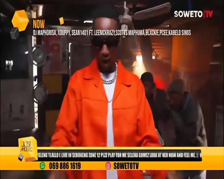 A man in an orange jumpsuit, shades, and a chain is center stage, performing on Soweto TV. Behind him, figures move in the dimly lit space, while the screen displays the "Just Music" logo.

