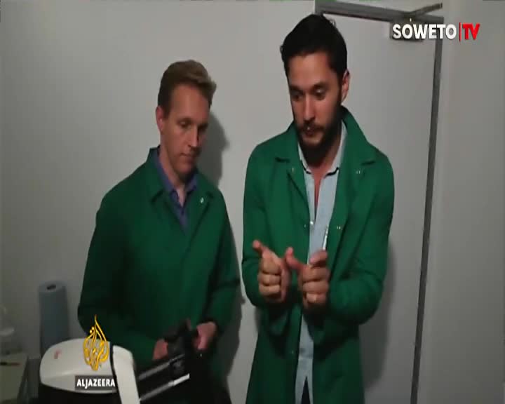 Two men in green lab coats stand near a piece of equipment, one gesturing with his hands. A Soweto TV logo appears in the upper right corner.