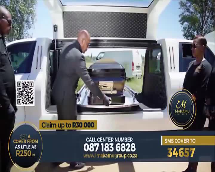 A man in a grey suit carefully slides a dark coffin with gold trim into the back of a white hearse. Another man in a black shirt stands by the open door, watching.