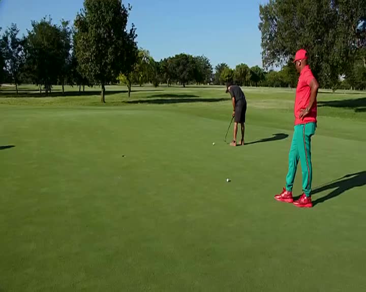The man in the red shirt and green pants stands with his hands on his hips, watching the other golfer line up his putt. The golfer in black bends low, ready to strike the ball on the bright green Soweto course.