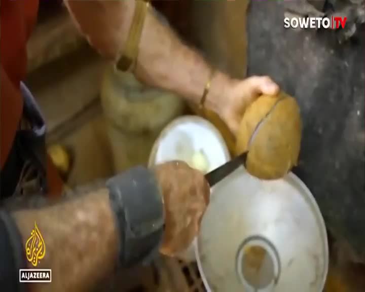 A man's hands slice a coconut in half over a metal bowl. This is happening in a busy Soweto market, likely for a Soweto TV segment.