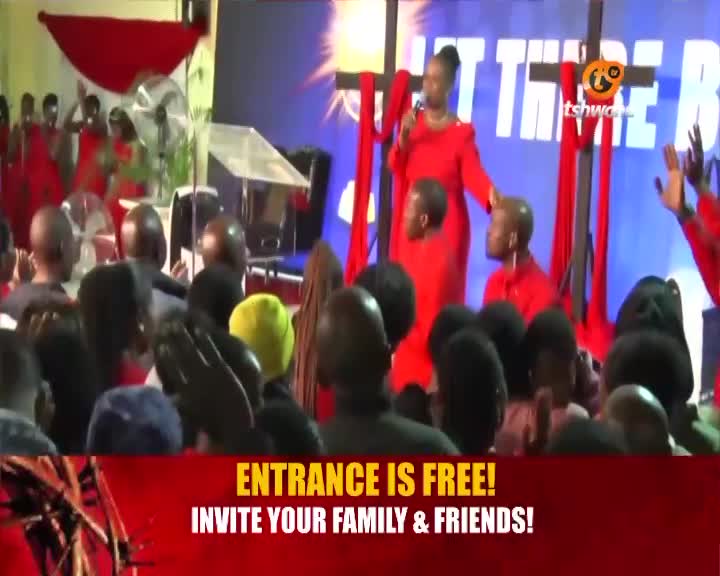 A woman in a bright red dress holds a microphone, speaking to a crowd gathered in a hall. The Tshwane TV logo is visible on a screen behind her, and a banner at the bottom announces "ENTRANCE IS FREE!" A woman in a bright red dress holds a microphone, speaking to a crowd gathered in a hall. The Tshwane TV logo is visible on a screen behind her, and a banner at the bottom announces "ENTRANCE IS FREE!"
