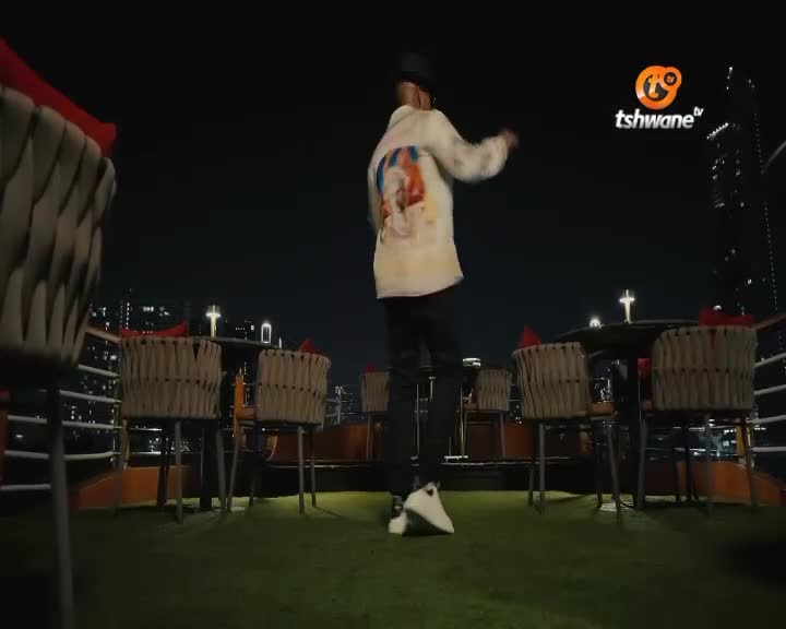 A person in a light-colored jacket with a graphic on the back dances on a rooftop overlooking Tshwane at night. The Tshwane TV logo glows brightly in the dark sky. A person in a light-colored jacket with a graphic on the back dances on a rooftop overlooking Tshwane at night. The Tshwane TV logo glows brightly in the dark sky.