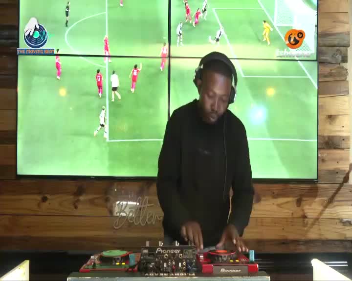 The DJ spins tracks, his hands moving across the Pioneer decks while a soccer match plays out on the screens behind him. The game unfolds on a bright green pitch, with players in red and white kits running across the field. The DJ spins tracks, his hands moving across the Pioneer decks while a soccer match plays out on the screens behind him. The game unfolds on a bright green pitch, with players in red and white kits running across the field.