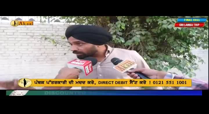 A man in a black turban is being interviewed, holding a microphone from Akaal Channel.  The microphones are pointed towards his face, with a brick wall and foliage in the background.
