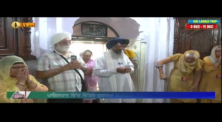 A man with a long white beard, wearing a white turban, is holding a paper and speaking. Several people are gathered, some with hands clasped in prayer, while others are using their phones.
