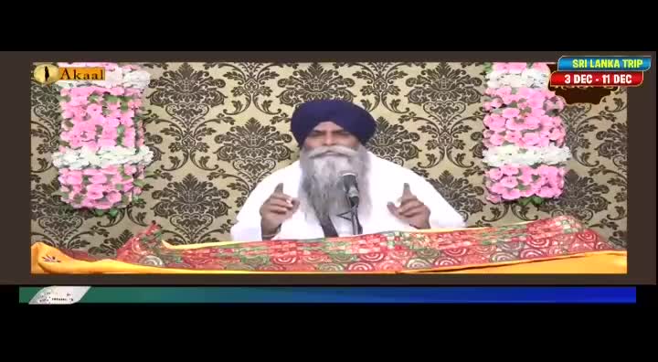 A man with a long white beard and a blue turban speaks into a microphone, gesturing with his hands. Behind him, the backdrop is a patterned wallpaper, and the Akaal Channel logo is visible in the corner.
