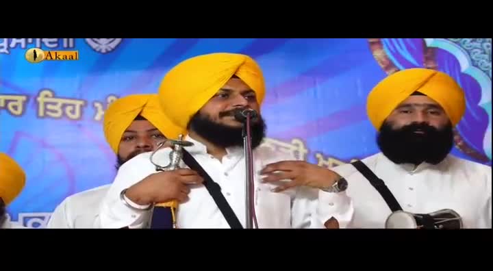 A man in a yellow turban speaks into a microphone, holding a ceremonial sword. Other men in similar attire stand beside him on a stage, likely for a broadcast on Akaal Channel from the United Kingdom.
