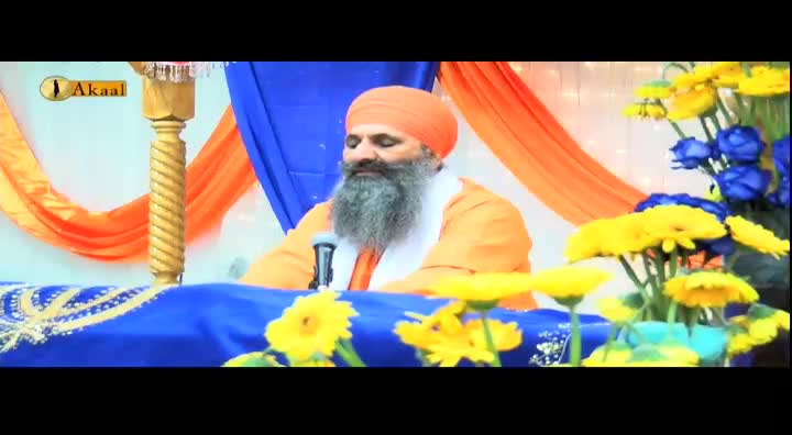 A man in an orange turban and beard sits at a table, speaking into a microphone. The Akaal Channel logo is visible, and the backdrop is decorated with flowers and draped fabric.
