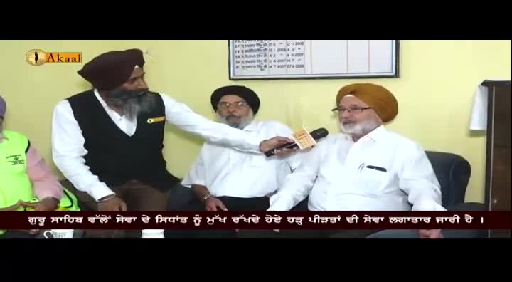 A man in a dark turban holds a microphone toward another man wearing a yellow turban. The Akaal Channel logo is visible in the corner.
