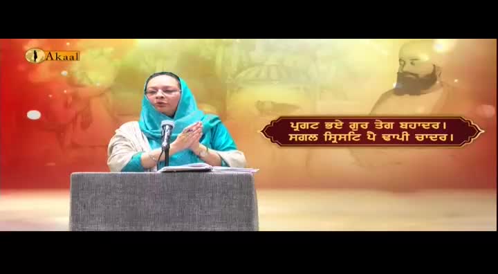 A woman in a teal head covering speaks into a microphone, her hands clasped in front of her. The Akaal Channel logo is visible in the corner, and a religious figure is in the background.
