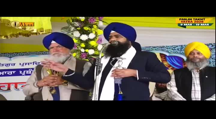 A man in a navy suit and turban speaks into a microphone, holding a ceremonial sword. Two other men, also wearing turbans, listen attentively on either side of him.
