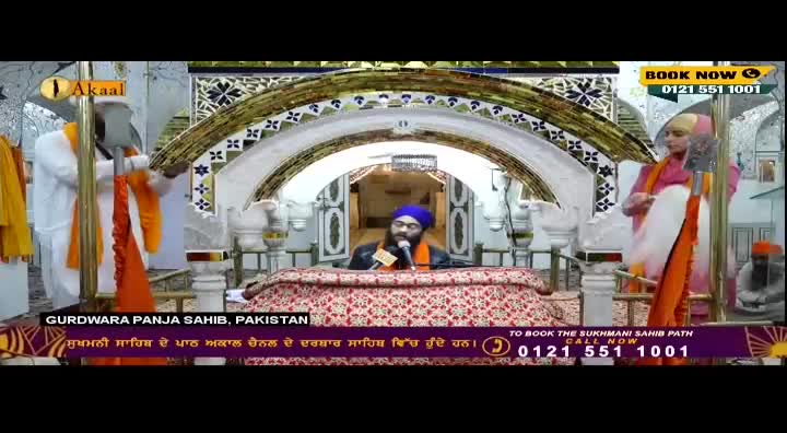A man in a blue turban speaks into a microphone from behind a decorated table, while others stand nearby, holding orange and white cloth. The scene is being broadcast on Akaal Channel, with a phone number displayed at the bottom of the screen.
