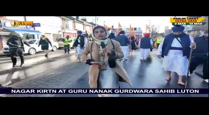 A woman from Akaal Channel is reporting live from a procession in Luton, United Kingdom. People in traditional attire are walking down the street, some barefoot, with flags and banners.
