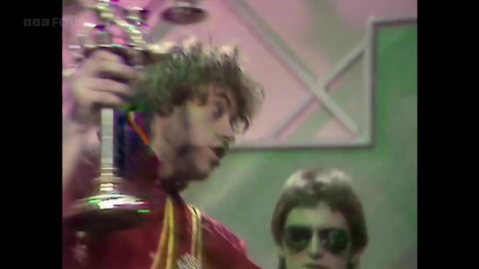 A man with curly hair, wearing a red jacket, holds a shiny object aloft. Another man, wearing sunglasses, stands behind him.