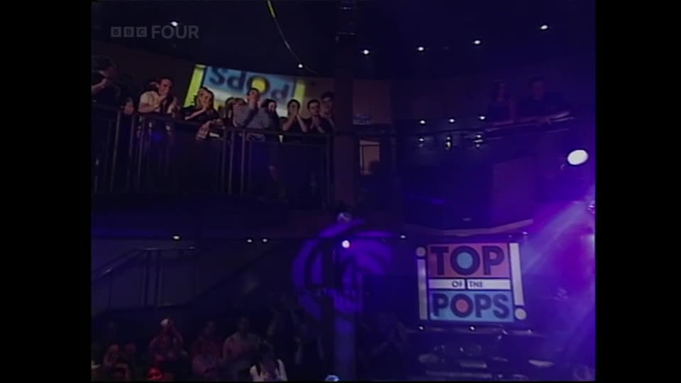A crowd is gathered on a balcony, looking down at the stage. The iconic "Top of the Pops" logo glows under the purple lights.