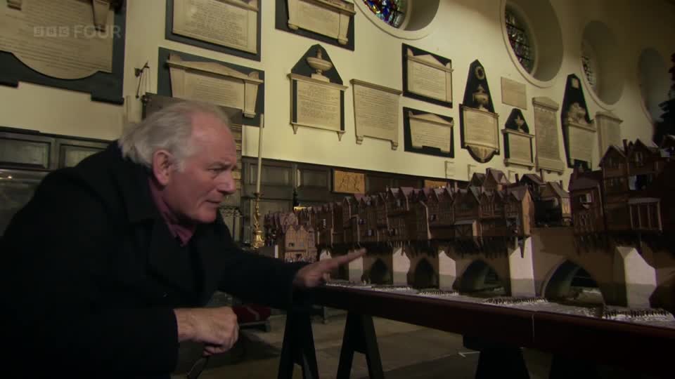 A man gestures towards a detailed model of houses built over a bridge, a scene from a BBC Four programme. The walls behind him are lined with framed plaques.