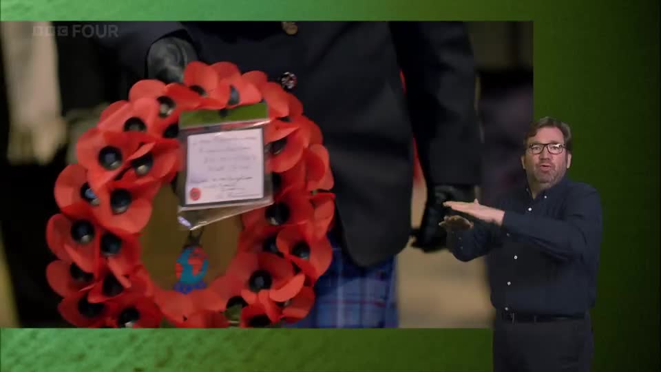 A man in a dark shirt gestures with his hands as a wreath of red poppies is presented. The BBC Four logo appears in the corner of the screen.
