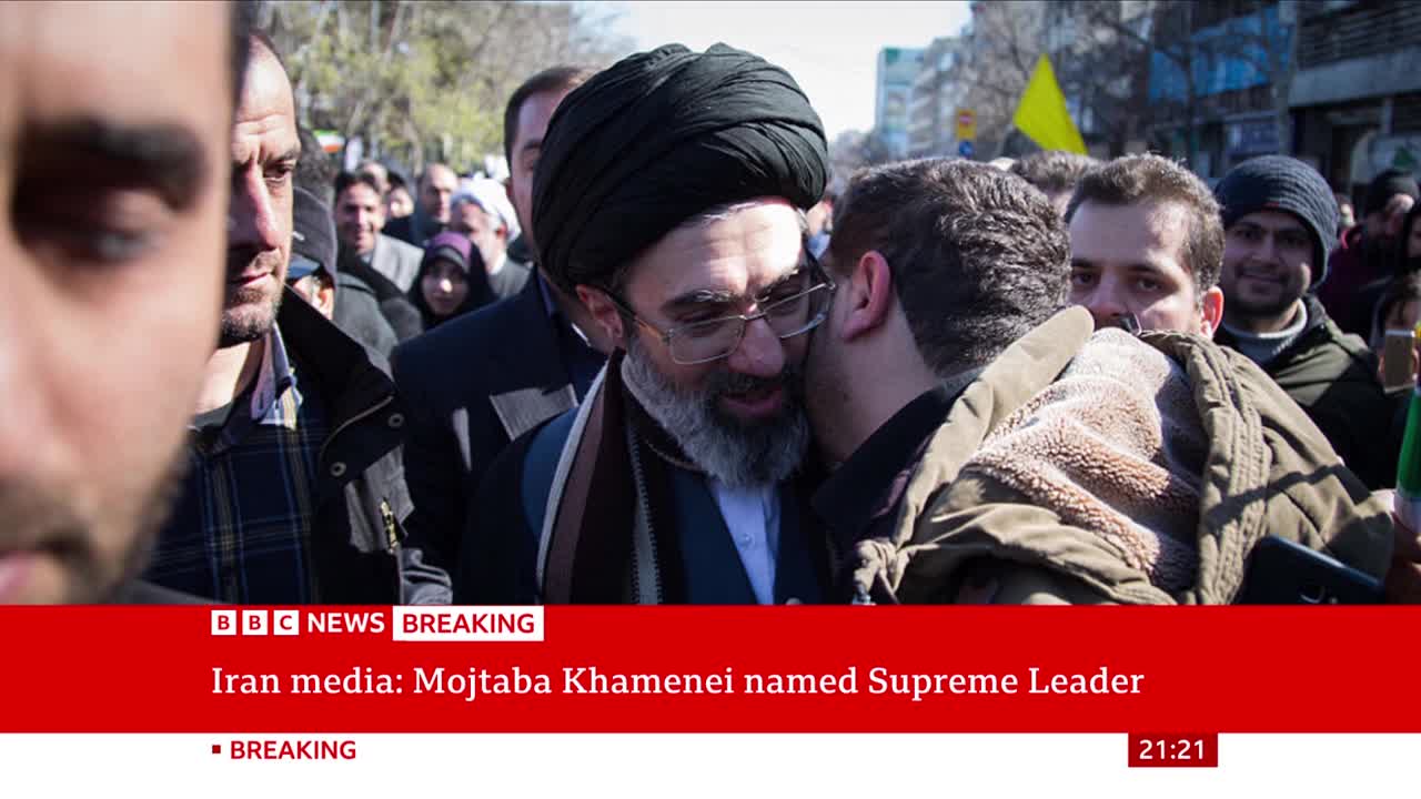 A man in a black turban leans in as another man whispers to him. A crowd presses in around them, some faces turned towards the camera. A man in a black turban leans in as another man whispers to him. A crowd presses in around them, some faces turned towards the camera.