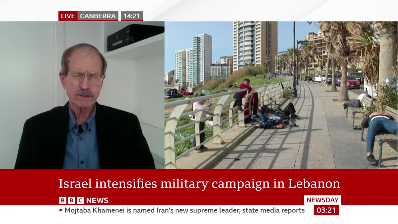 People are resting on benches and the ground along a waterfront promenade. Tall buildings and palm trees line the street behind them. People are resting on benches and the ground along a waterfront promenade. Tall buildings and palm trees line the street behind them.