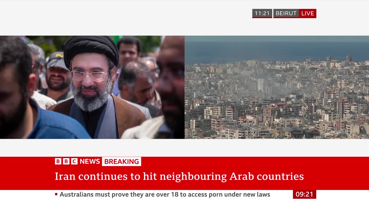 A man in a black turban and glasses smiles as he's surrounded by others. Beyond him, a dense city stretches towards the sea under a hazy sky. A man in a black turban and glasses smiles as he's surrounded by others. Beyond him, a dense city stretches towards the sea under a hazy sky.