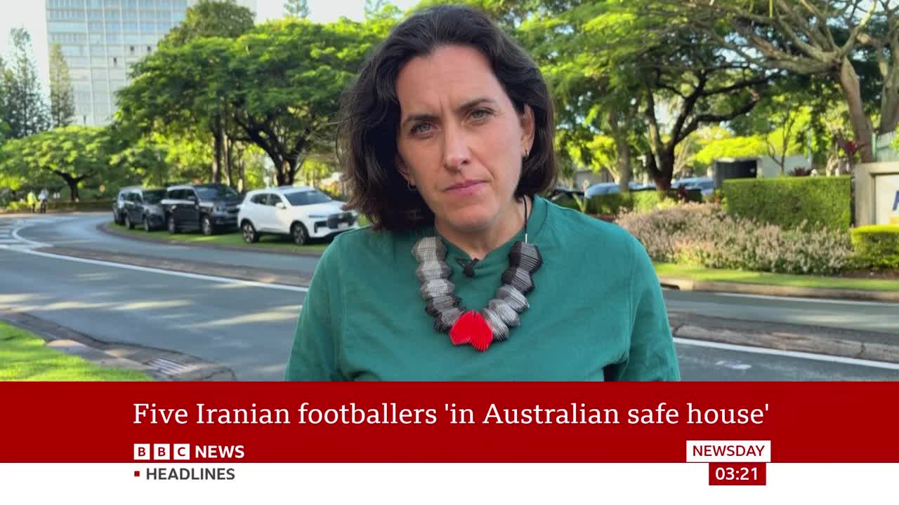 A woman stands on a street, speaking to the camera. Cars are parked on the side of the road behind her. A woman stands on a street, speaking to the camera. Cars are parked on the side of the road behind her.