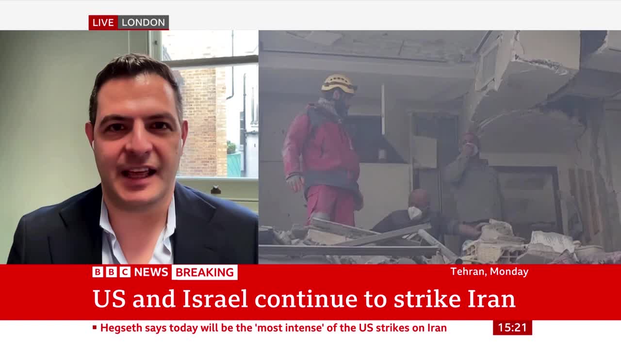 A man in a red jacket and hard hat stands amidst rubble, looking towards the right. Another person, wearing a mask, is visible in the background, also surrounded by debris. A man in a red jacket and hard hat stands amidst rubble, looking towards the right. Another person, wearing a mask, is visible in the background, also surrounded by debris.