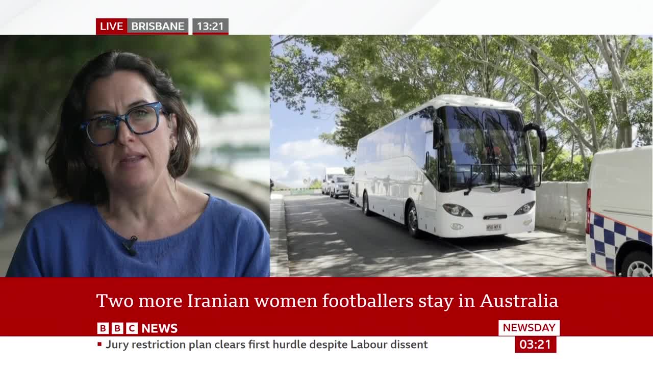 A line of white buses and vans moves slowly down a road lined with trees. A police vehicle with a distinctive blue and white checkered pattern is visible on the right. A line of white buses and vans moves slowly down a road lined with trees. A police vehicle with a distinctive blue and white checkered pattern is visible on the right.